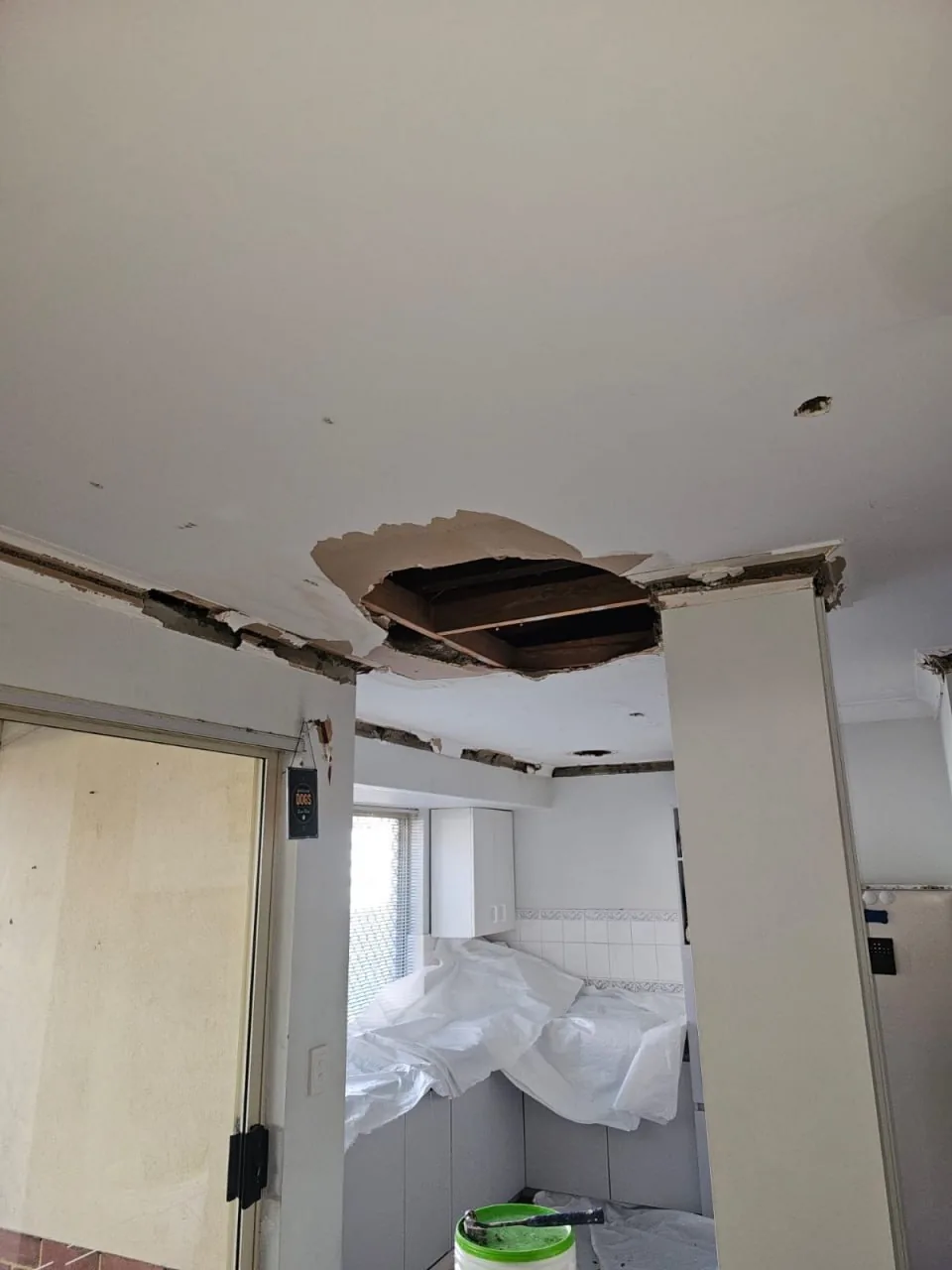 A section cut out of a Bassendean ceiling exposing the timber battens above, with the cornice stripped along the wall line and dust sheets over the kitchen below