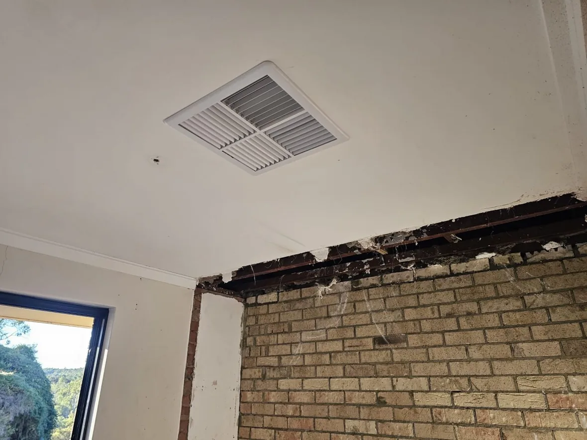 A Roleystone ceiling cut back around a ceiling vent to expose the framing above it, with the brick wall and window alongside