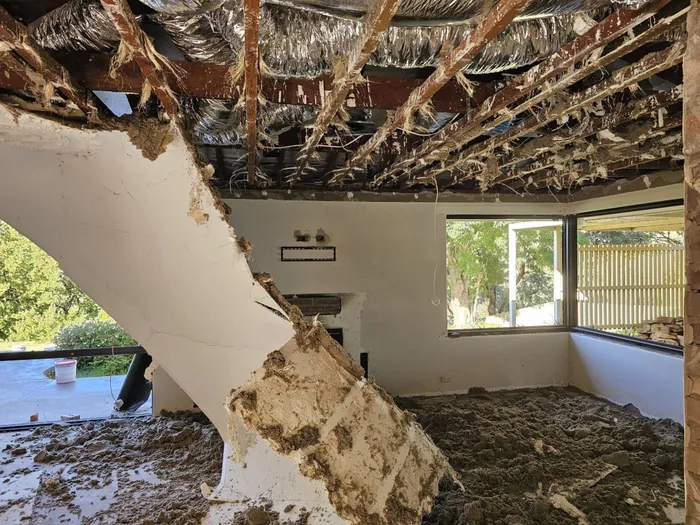 A whole sheet of old plasterboard ceiling folded down into a Roleystone room with debris across the floor below it, the roof timbers exposed above and windows looking out to the garden